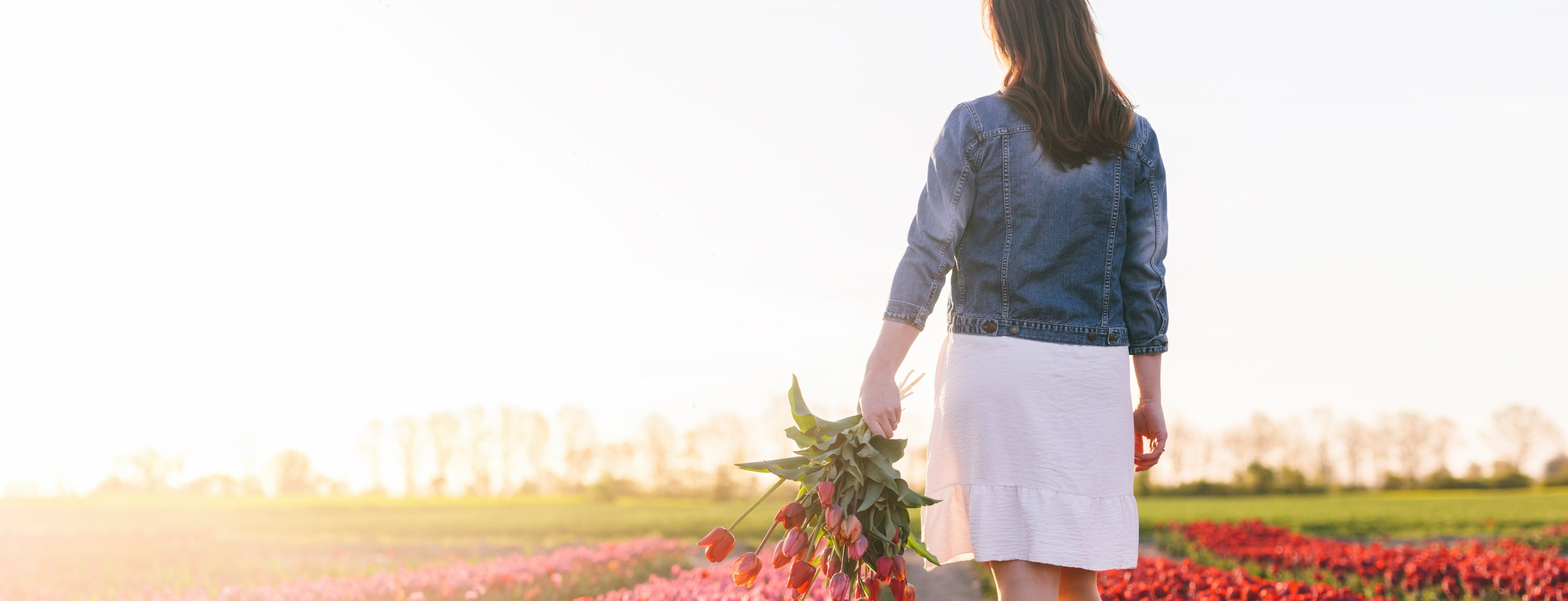 Mujer caminando por un campo de flores al atardecer, sosteniendo un ramo de flores, en un entorno natural y luminoso.