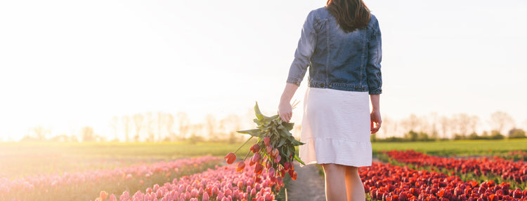 Mujer caminando por un campo de flores al atardecer, sosteniendo un ramo de flores, en un entorno natural y luminoso.